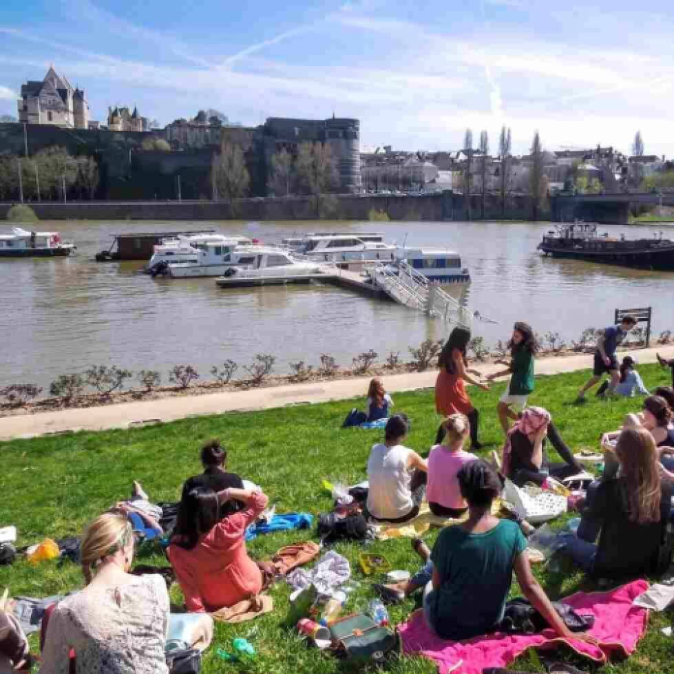 Group of study abroad students sit in the grass on a sunny day alongside a canal while boats ride by.